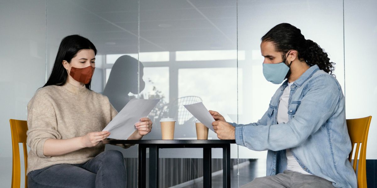 Two people wearing face masks sit at a table reviewing papers; one has curly hair, the other wears a sweater
