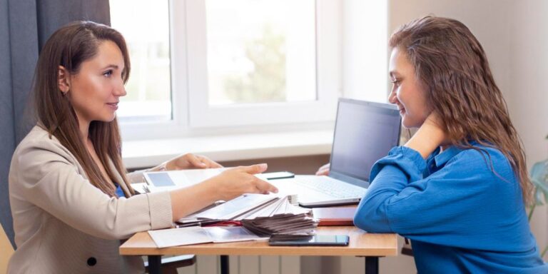 Two women are seated across a table, one reviewing papers with the other in an office setting