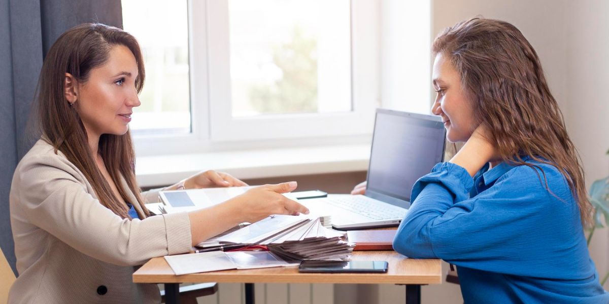 Two women are seated across a table, one reviewing papers with the other in an office setting