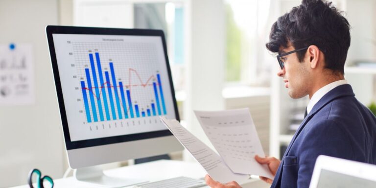 Man in a suit and glasses reviewing papers at a desk with a monitor displaying a bar and line graph