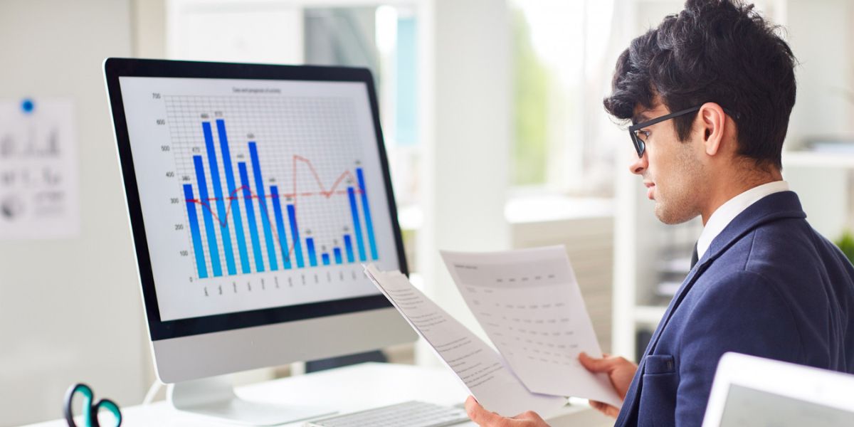 Man in a suit and glasses reviewing papers at a desk with a monitor displaying a bar and line graph