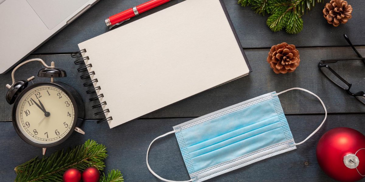 A medical mask, alarm clock, and blank notebook on a wooden desk with festive Christmas ornaments and pine branches.