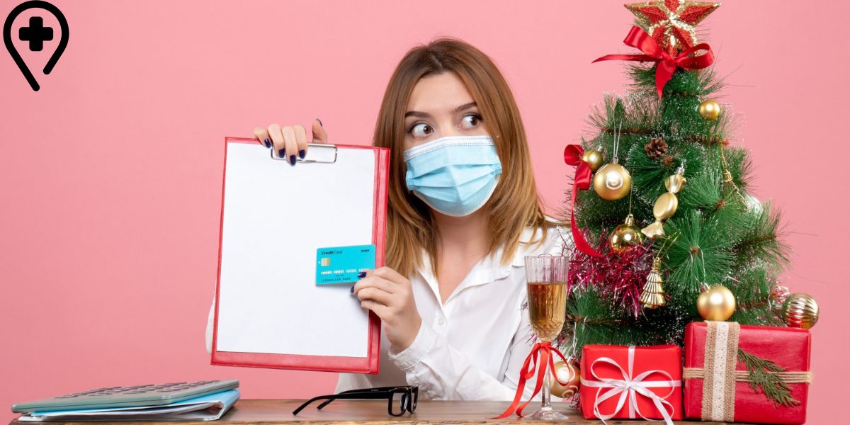 A masked nurse holds a clipboard and credit card next to a decorated Christmas tree with gifts and a glass of cider.