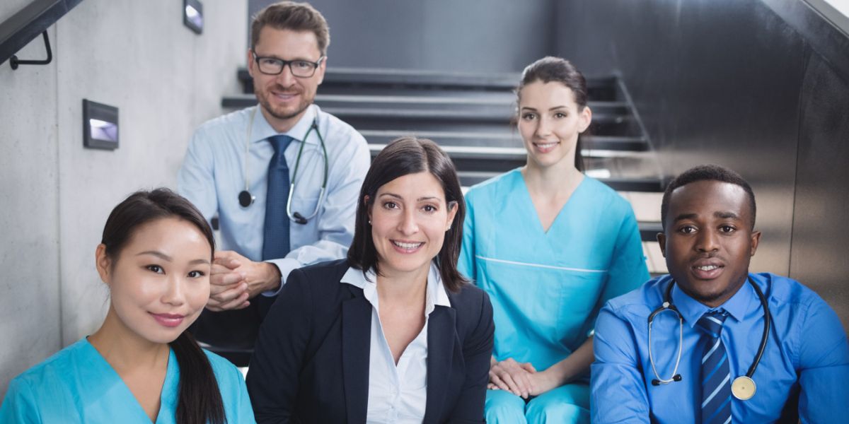 A diverse group of smiling medical professionals, including nurses and doctors, sitting together on office stairs